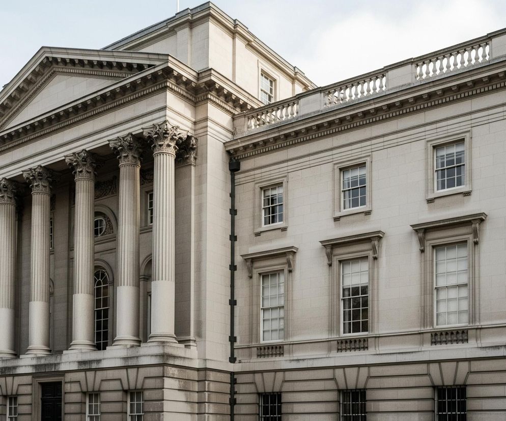 Jasnoszary budynek o klasycystycznej architekturze dominuje w centrum obrazu, prezentując symetryczną fasadę z dużą ilością detali. Główne wejście charakteryzuje się portykiem z sześcioma korynckimi kolumnami, wspierającymi trójkątny fronton z gzymsem. Pozostała część budynku posiada liczne prostokątne okna z białymi ramami i drobnymi szprosami, niektóre z nich ozdobione są naczółkami i ozdobnymi wspornikami. Na dachu widoczna jest balustrada z balaskami.