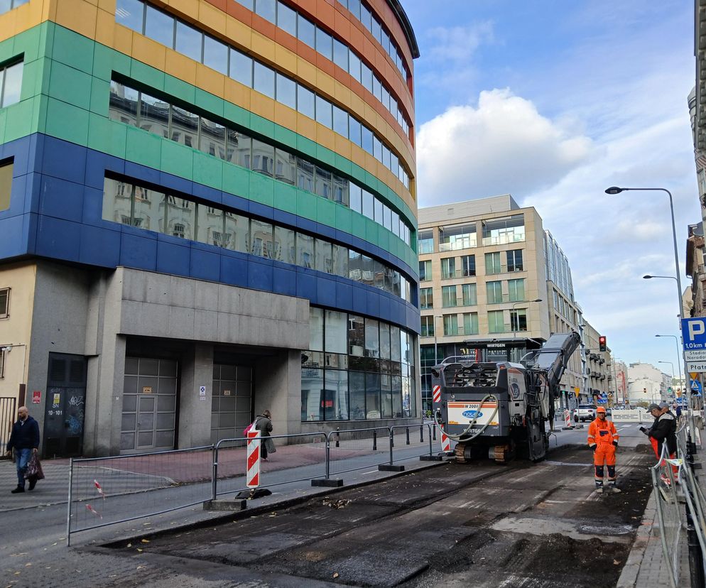 Ulica Okrzei w Warszawie w trakcie przebudowy, z widoczną frezarką drogową i robotnikami w pomarańczowych kombinezonach, obok nowoczesnego budynku o kolorowych fasadach i zabytkowej kamienicy. O tej zielonej metamorfozie przeczytasz na Architektura Murator Plus.