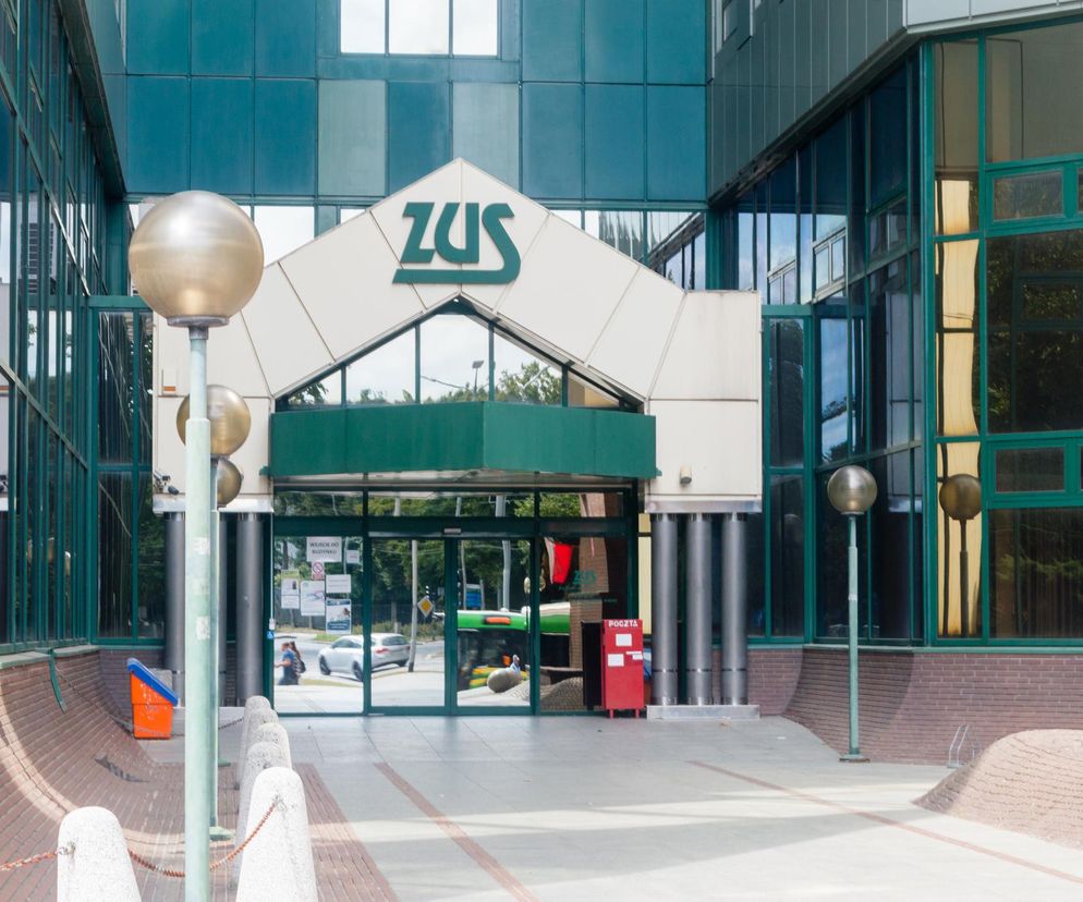 Front budynku ZUS z dużym, zielonym logo na dachu. W tle zielone przeszklenia i odbijające się światło. Przed wejściem do budynku widoczne lampy uliczne oraz czerwona skrzynka pocztowa. Zdjęcie ilustruje obciążenia ZUS dla przedsiębiorców, o których przeczytasz na Super Biznes.