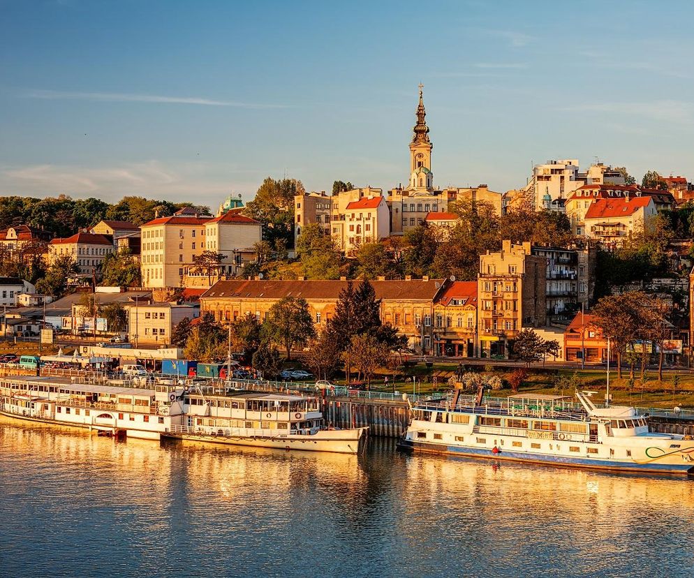 Widok na nabrzeże Sawy w Belgradzie o zachodzie słońca z budynkami i kościołem na wzgórzu, oraz łodziami zacumowanymi na rzece. To krajobraz architektoniczny, który może być inspiracją dla studentów konkursu Saint-Gobain, o którym przeczytasz na Architektura Murator Plus.