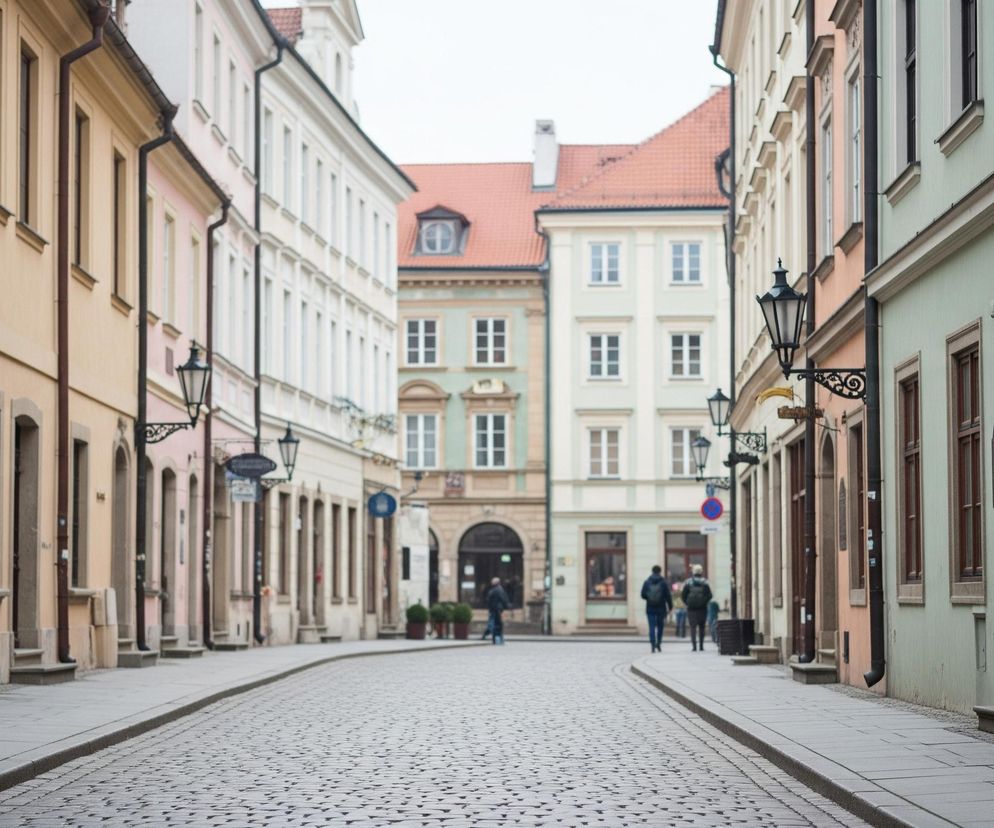 Wąska, brukowana ulica z chodnikami po obu stronach rozciąga się w perspektywie, otoczona kolorowymi kamienicami o pastelowych fasadach. Po prawej i lewej stronie widoczne są dwu- i trzypiętrowe budynki w odcieniach beżu, różu, jasnozielonego i kremowego, z licznymi brązowymi oknami i ozdobnymi latarniami ulicznymi wiszącymi na elewacjach. Na końcu ulicy, w tle, widoczny jest większy budynek z pomarańczowym dachem i lukarną, otoczony innymi kamienicami, a kilka niewyraźnych postaci w ciemnych ubraniach przechodzi przez ulicę.