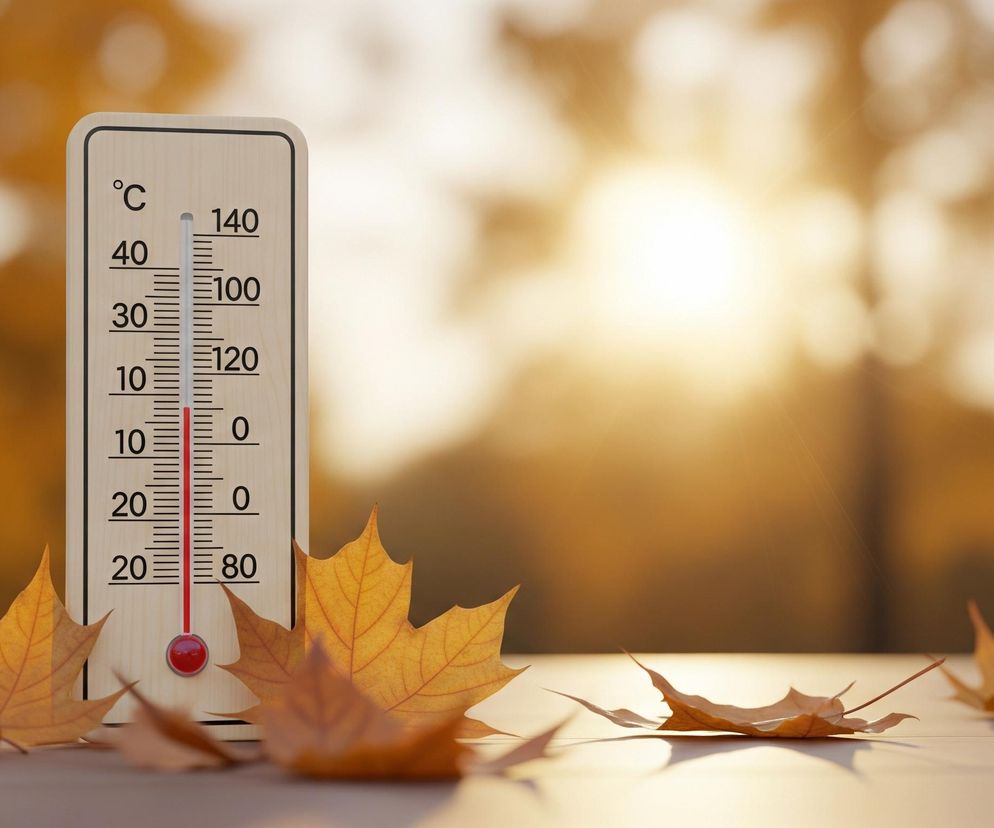 Centralnie na zdjęciu stoi beżowy termometr z czarnymi napisami i czerwoną skalą rtęciową. Rtęć wskazuje temperaturę około 15 stopni Celsjusza, z cyframi po lewej stronie od 40 do -20 w stopniach Celsjusza i po prawej od 140 do 80 w stopniach Fahrenheita. U podstawy termometru oraz na jasnej drewnianej powierzchni, na której stoi, rozrzucone są pomarańczowo-brązowe liście jesienne. Tło jest rozmyte, dominują ciepłe odcienie pomarańczu i brązu, z wyraźnym, jasnym światłem w centrum, prawdopodobnie słońcem, tworzącym miękki blask.