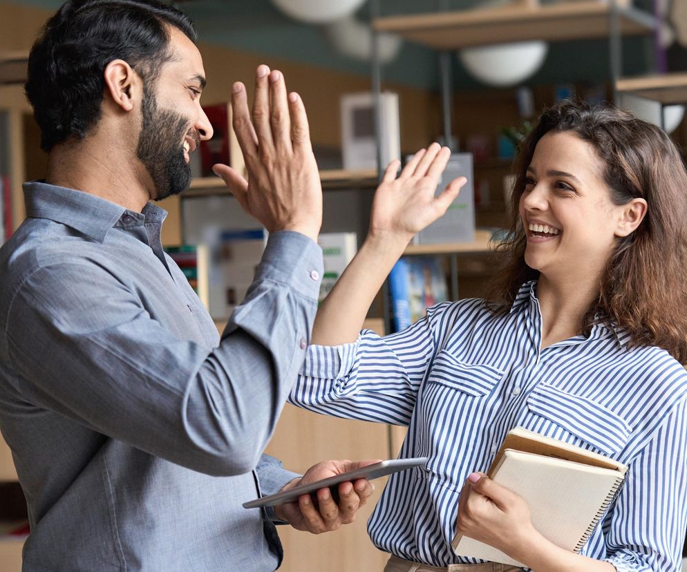 Dwoje uśmiechniętych ludzi, kobieta i mężczyzna, wymieniających high five w biurze. Mężczyzna trzyma tablet, kobieta notatniki. Obraz symbolizuje sukces, współpracę i zadowolenie z pracy, idealnie odzwierciedlając pozytywne zmiany w zatrudnieniu, o których przeczytasz na Super Biznes.