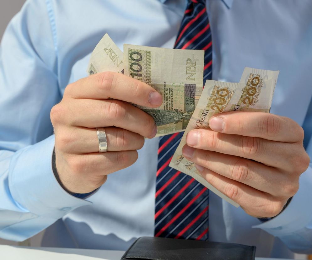 Mężczyzna w błękitnej koszuli i krawacie liczy banknoty 100 i 200 złotych, trzymając je w dłoniach nad portfelem. Symbolizuje to finanse i bezpieczeństwo pieniędzy, o którym można przeczytać więcej na portalu Super Biznes w kontekście incydentu z Santanderem.