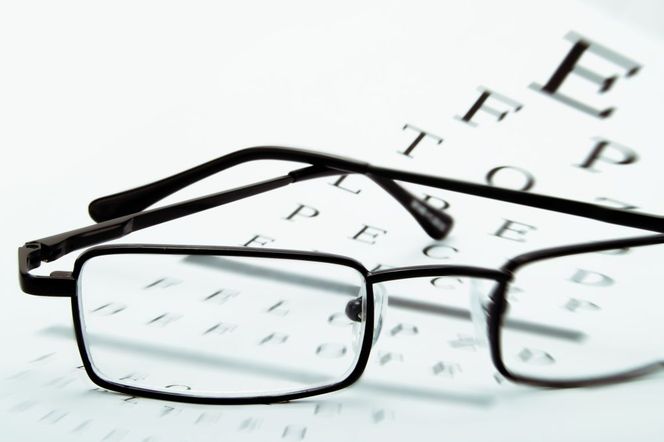 Czarne okulary leżące na tablicy Snellena do badania wzroku, co symbolizuje potrzebę korekcji presbiopii, czyli starczowzroczności. Więcej o tej wadzie wzroku i metodach jej leczenia znajdziesz na Poradnik Zdrowie.