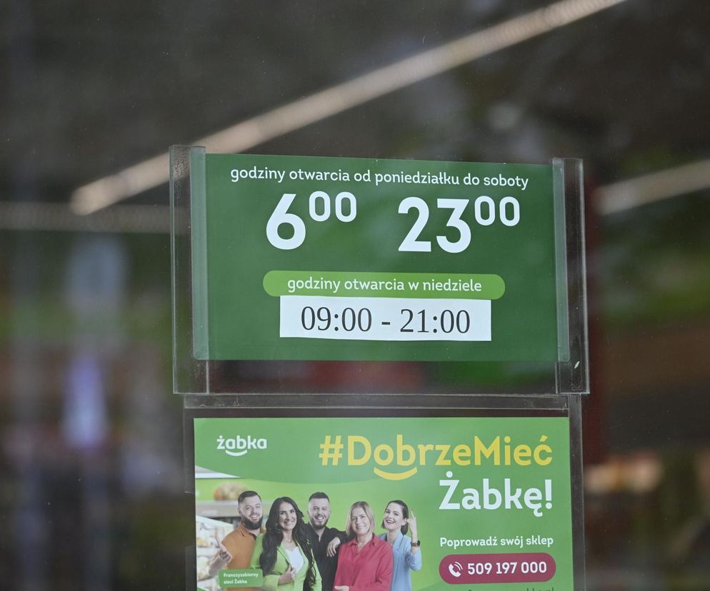 Tablica z godzinami otwarcia sklepu Żabka: poniedziałek-sobota 6:00-23:00, niedziele 9:00-21:00. Poniżej widoczny baner reklamowy z hasłem #DobrzeMiećŻabkę! To przykład sklepu otwartego w niedziele niehandlowe, o czym można przeczytać na Super Biznes.