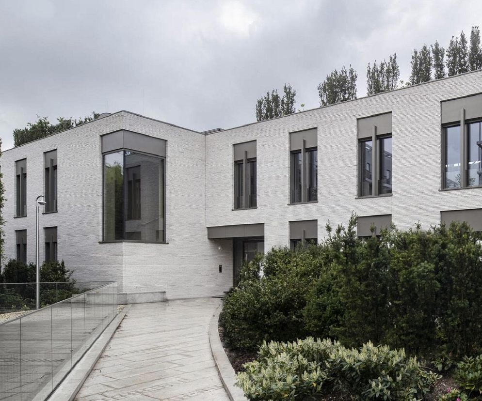 Biała Willa w Warszawie, nowoczesny apartamentowiec z jasnej cegły z dużymi oknami i zielonymi tarasami na dachu, otoczony bujną roślinnością. Budynek o modernistycznej formie, łączący elegancję z funkcjonalnością, idealnie wpisuje się w ideę willi miejskiej, o której możesz przeczytać na Architektura Murator Plus.