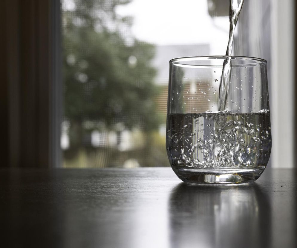 Woda z kranu w szklance stojącej na ciemnym blacie, w tle rozmazane okno z widokiem na zieleń. Strumień wody tworzy bąbelki, symbolizując dbałość o zdrowie i alternatywę dla wody butelkowanej, o której szkodliwości przeczytasz na Poradnik Zdrowie.