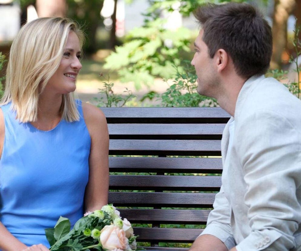 Aktorzy Marta Chodorowska jako Julia i Michaił Pszeniczny jako Dima, siedzący na ławce. Julia w niebieskiej sukience, z bukietem kwiatów, uśmiecha się do Dimy. Oboje patrzą na siebie, ich gesty sugerują rozmowę o ciąży Julii i nadchodzącym ślubie w serialu M jak miłość. Więcej na Super Seriale.