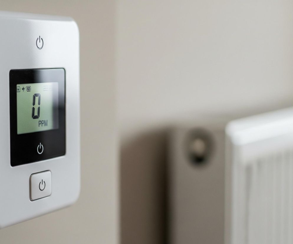 Na beżowej ścianie, po lewej stronie kadru, zamontowane jest białe, prostokątne urządzenie z zaokrąglonymi rogami. Posiada ono czarny wyświetlacz z zielonym podświetleniem, na którym widać dużą cyfrę 0 oraz tekst PPM poniżej, a także ikony baterii i sygnału w górnej części. Powyżej wyświetlacza znajduje się wytłoczony symbol włącznika, podobny symbol widać poniżej ekranu i na fizycznym, kwadratowym przycisku zlokalizowanym na samym dole urządzenia. W tle, po prawej stronie, rozmyty jest biały grzejnik żeberkowy.