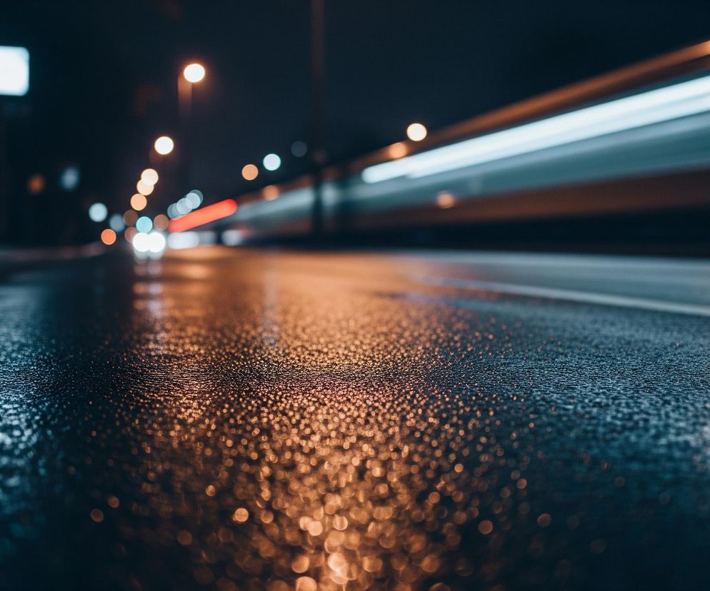 Mokry, asfaltowy chodnik pokryty jest drobnymi kroplami wody, które odbijają światła otoczenia, tworząc rozproszone, pomarańczowo-niebieskie refleksy bokeh. W tle widoczna jest rozmyta ulica nocą, po której przemieszcza się pojazd, pozostawiając za sobą wydłużone smugi jasnego, białego światła. Po lewej stronie ulicy, również w tle, dostrzegalne są rozmyte, okrągłe światła ulicznych latarni w odcieniach pomarańczy i bieli. Całość zanurzona jest w ciemnej, granatowej tonacji nocy, z wyraźnym kontrastem między ostrością kropli wody na pierwszym planie a rozmyciem dalszych elementów.