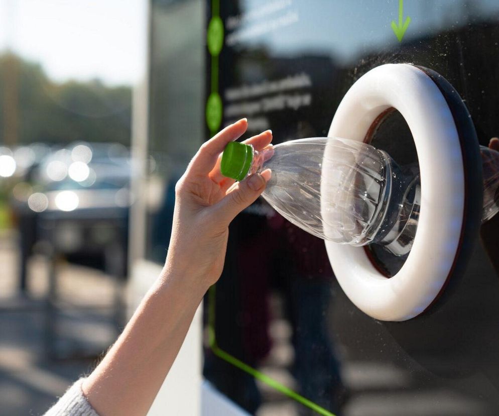 Dłoń osoby wkładającej plastikową butelkę PET z zieloną nakrętką do otworu butelkomatu. Widoczny element systemu kaucyjnego, który usprawni recykling opakowań, o czym przeczytasz na portalu Super Biznes. W tle niewyraźne zarysy samochodów na parkingu.