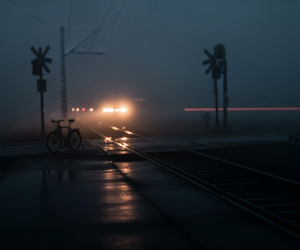 Przedstawiono scenę kolejową w warunkach słabej widoczności, prawdopodobnie mgły lub zmierzchu. Na pierwszym planie po lewej stronie znajduje się sylwetka roweru, a obok niego, na środku obrazu, tory kolejowe, których powierzchnia odbija jasne światło nadjeżdżającego pociągu. Tory ciągną się w głąb obrazu, gdzie na horyzoncie widoczne są intensywnie świecące żółte reflektory pociągu, a także kilka mniejszych, okrągłych świateł, w tym czerwone. Po lewej i prawej stronie torów znajdują się słupy z sygnałami przejazdowymi w kształcie litery X, a po prawej stronie w tle rozciąga się długi, rozmyty czerwony pas światła. Całość utrzymana jest w ciemnych, błękitno-szarych tonacjach, z wyraźnymi kontrastami jasnych świateł.