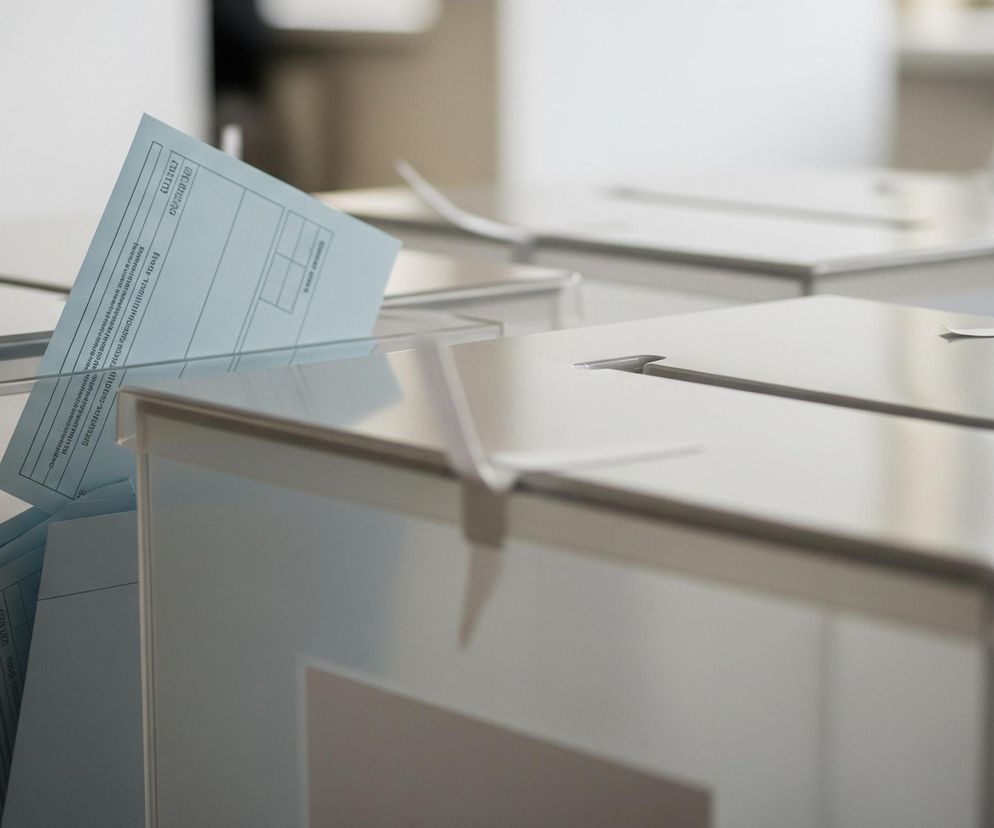 Na pierwszym planie, z prawej strony, znajduje się przezroczysta urna wyborcza z wpadającą do środka, jasnoniebieską kartą. Wewnątrz urny widać ułożone podobne, niebieskie karty. W tle widoczne są inne, częściowo zamazane, białe lub przezroczyste urny, z podłużnymi otworami na górze i białymi taśmami zabezpieczającymi na ich krawędziach. Ogólna kolorystyka jest jasna, dominują odcienie bieli, szarości i błękitu.