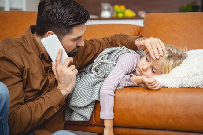 Zmartwiony ojciec trzyma telefon przy uchu i sprawdza czoło chorej córki, która leży na kanapie pod kocem. Obraz symbolizuje troskę rodziców o zdrowie dzieci oraz zagrożenie poważnymi powikłaniami po ponownym zakażeniu COVID-19. Więcej na ten temat przeczytasz na Poradnik Zdrowie.