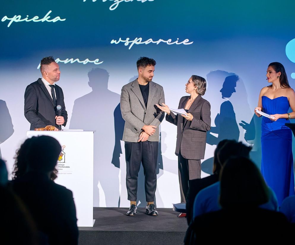 Aukcja ART&Charity Fundacji Ronalda McDonalda: Anna Dereszowska w niebieskiej sukni i Marcin Wójcik prowadzą wydarzenie. Widoczni również kurator Michał Borowik i inni prelegenci na tle ekranu z napisami opieka, wsparcie, pomoc. O wydarzeniu przeczytasz na Super Biznes.