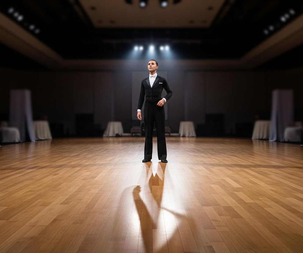 Młody mężczyzna w eleganckim czarnym fraku i białej muszce stoi centralnie na błyszczącym, drewnianym parkiecie. Jego wzrok skierowany jest lekko w górę, a lewa dłoń swobodnie spoczywa w kieszeni spodni, podczas gdy prawa ręka jest opuszczona wzdłuż tułowia. Postać jest oświetlona silnym, jasnym światłem z góry, tworzącym długi, rozmazany cień na podłodze, który rozciąga się w lewo i do przodu. Tło sali jest zaciemnione, z widocznymi, niewyraźnymi elementami architektonicznymi i białymi, udrapowanymi tkaninami po bokach, oraz kilkoma punktowymi światłami na suficie.