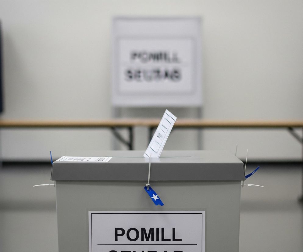 Na pierwszym planie, centralnie, stoi szara urna wyborcza z włożoną do szczeliny białą kartą. Urna jest zabezpieczona niebieskimi opaskami zaciskowymi, do których przymocowane są niebieskie pieczęcie z białą gwiazdką. Na froncie urny widnieje czarno-biała etykieta z napisem POMILL SEAD. W tle, po lewej i prawej stronie, widać ciemnoniebieskie, rozmyte kabiny do głosowania, a za urną znajduje się długi drewniany stół i kolejny, podobny plakat z tekstem POMILL SEAD. Pomieszczenie ma jasnoszare ściany i ciemniejszą podłogę.