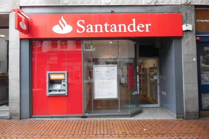 Fasada oddziału banku Santander z czerwonym szyldem i bankomatem. Zdjęcie ilustruje artykuł o metodach oszustów w kontekście skimmingu i ochrony przed kradzieżą danych z kart, o czym można przeczytać na portalu Super Biznes.