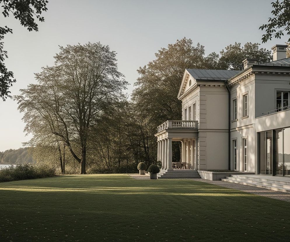 W centrum kadru widoczny jest duży, beżowy budynek w stylu klasycznym z nowoczesnymi elementami, położony w zielonym otoczeniu. Lewą stronę frontowej fasady zdobi portyk z czterema kolumnami, wspierający balkon z kamienną balustradą, a dalej na prawo rozciągają się duże, szklane panele okien od parteru do piętra. Przed budynkiem rozpościera się zadbany trawnik o intensywnie zielonym kolorze, częściowo oświetlony promieniami słońca, tworzącymi długie cienie. W tle za budynkiem oraz po lewej stronie kadru rozciąga się gęsty las liściasty, a w oddali po lewej stronie widoczne jest spokojne jezioro, otoczone również drzewami.