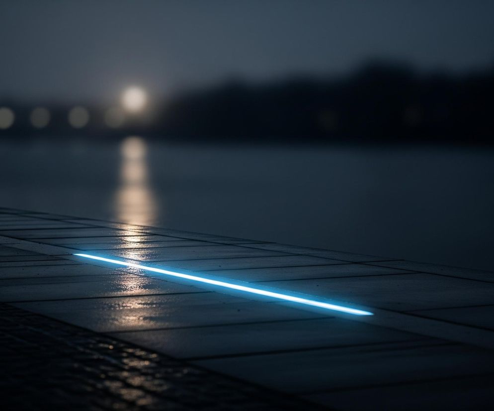 Ciemną scenę nocną, gdzie głównym elementem jest jasna, turkusowa linia świetlna biegnąca ukośnie od lewego dolnego rogu w kierunku prawego środka. Linia ta jest osadzona w mokrym, ciemnym chodniku złożonym z prostokątnych płyt. Powierzchnia chodnika odbija pomarańczowo-żółte światło z rozmazanych źródeł znajdujących się poza kadrem, tworząc refleksy na mokrej nawierzchni. W tle widoczny jest duży zbiornik wodny, a za nim niewyraźne, rozmyte światła w oddali oraz ciemne, nieostre sylwetki drzew lub budynków.