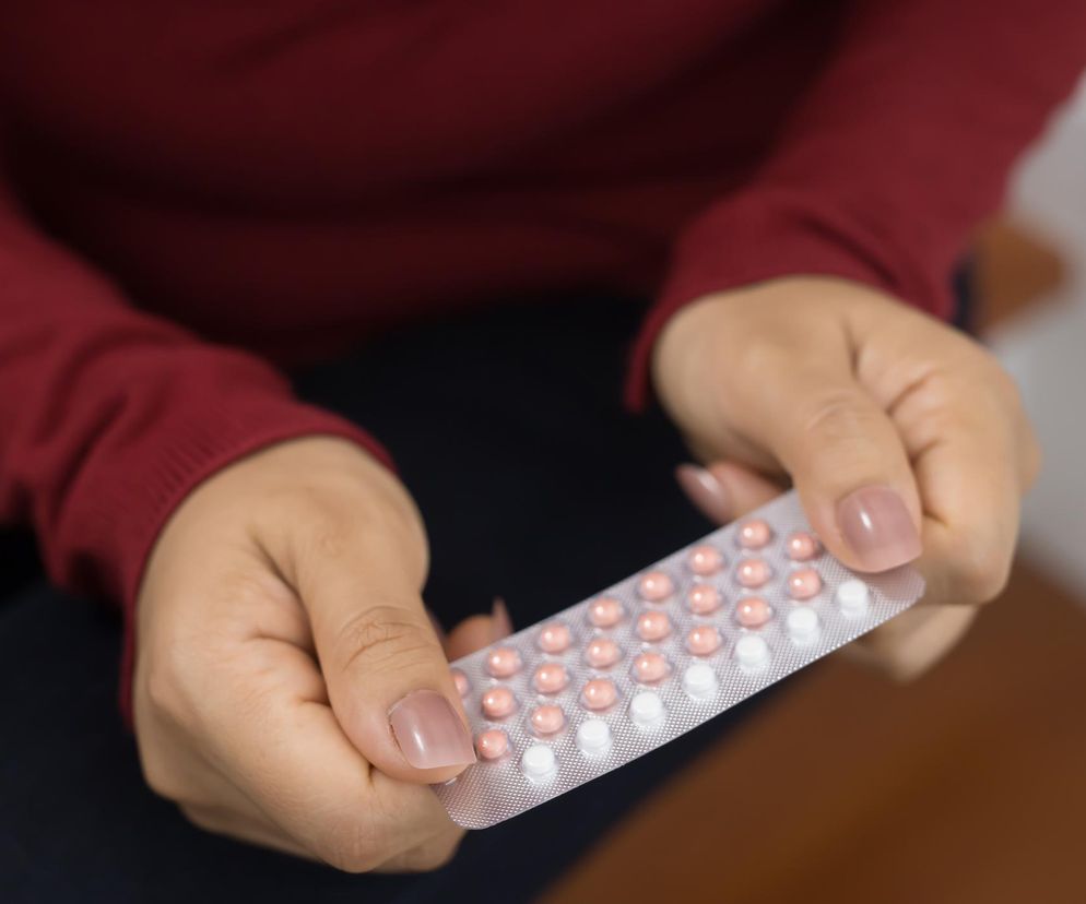 Kobieta w czerwonym swetrze trzyma w dłoniach blister tabletek antykoncepcyjnych, w rzędach ułożone są jasnoróżowe i białe pigułki. Dowiedz się, które składniki antykoncepcji hormonalnej mogą zwiększać ryzyko raka piersi, czytając na Poradnik Zdrowie.