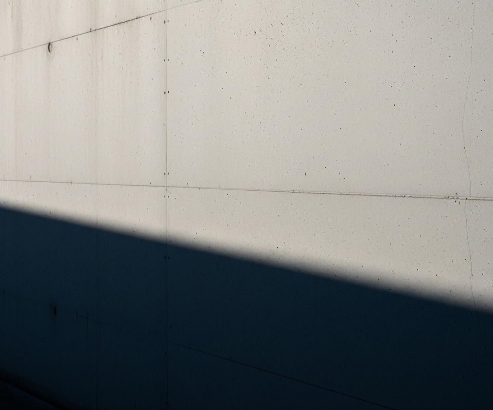Pionowa, jasnoszara ściana z betonu o wyraźnej teksturze, pokryta jest drobnymi ciemnymi kropkami i przebarwieniami. Przez całą szerokość ściany przebiega pozioma linia łącząca pionowe spoiny płyt, a w prawym górnym rogu widoczny jest okrągły element. Na lewej części ściany, od dołu do połowy wysokości, rzuca się wyraźny, ciemnoniebieski cień, oddzielony od jasnej części ostrą, ukośną linią.