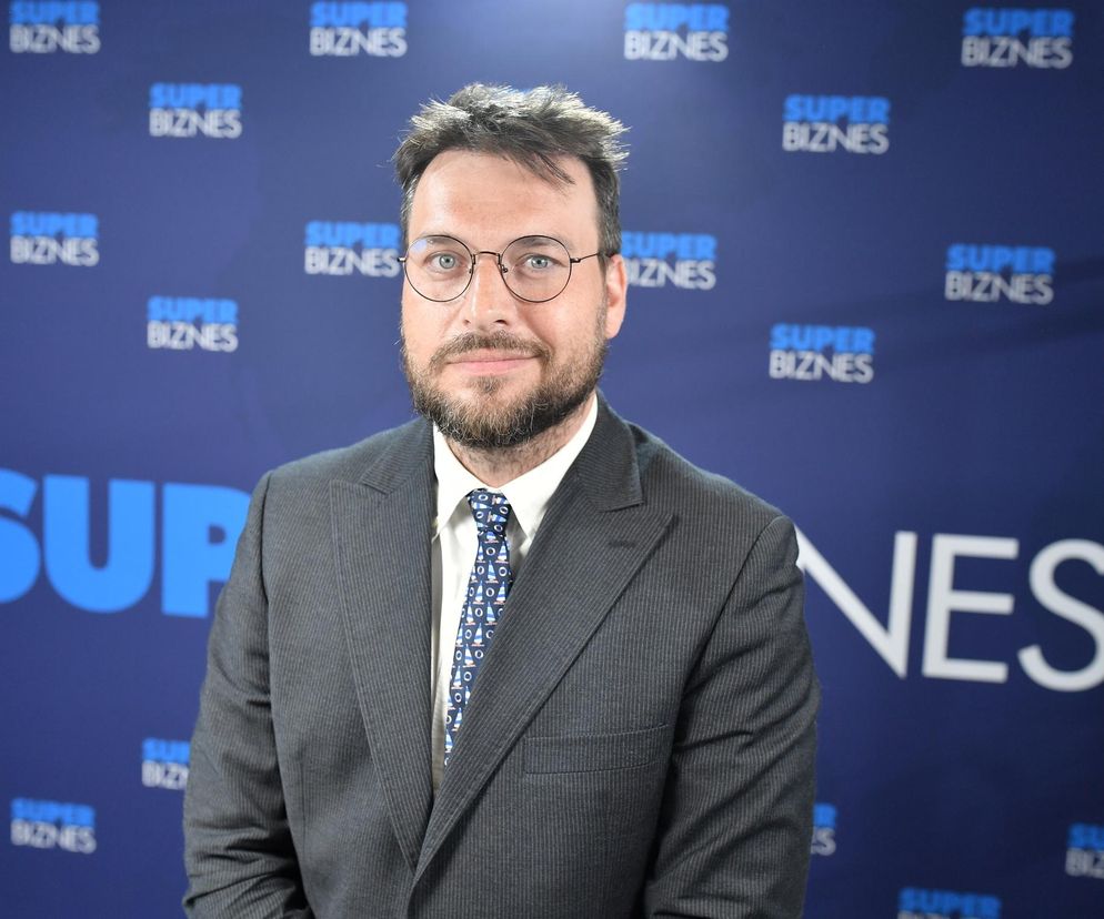 Piotr Arak, główny ekonomista VeloBanku, w okrągłych okularach, z brodą, w eleganckim garniturze i krawacie w białe żaglówki, stoi na tle niebieskiej ściany z logo Super Biznes, podsumowując rok 2025 i prognozując przyszłość gospodarczą, o czym przeczytasz na Super Biznes.