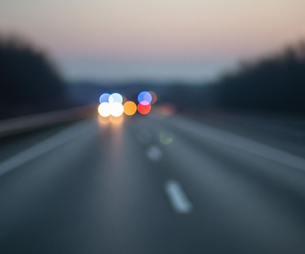 Na pierwszym planie widoczna jest szeroka, wielopasmowa droga asfaltowa z białymi liniami, ukazana w perspektywie, rozciągająca się ku horyzontowi. Po obu stronach drogi, w oddali, rozciągają się ciemne, niewyraźne sylwetki lasu lub drzew. Na drodze, w dali, znajdują się rozmyte światła, tworzące okrągłe plamy koloru białego, żółtego, niebieskiego i czerwonego, z dominującymi odcieniami niebieskiego i czerwonego. Całość obrazu jest nieostra, co tworzy efekt bokeh, szczególnie widoczny na światłach i elementach tła, z niebem w odcieniach szarości i lekkiego różu w tle.