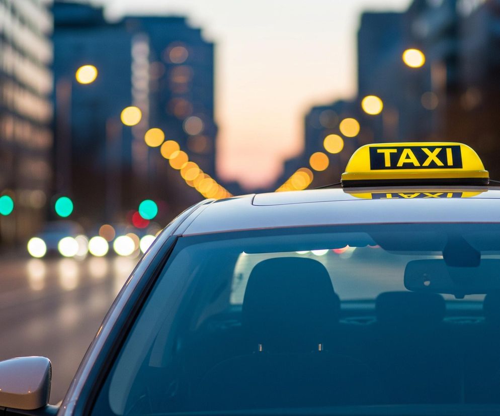 Przedni fragment samochodu z jasnym, żółtym szyldem TAXI na dachu, wyróżnia się na tle rozmytego, miejskiego krajobrazu. Na szyldzie, umieszczonym centralnie u góry kadru, widnieje czarny napis TAXI, a jego jaskrawe światło odbija się na szybie przedniej pojazdu. Wnętrze samochodu jest ciemne i niewyraźne, zarysowane są jedynie ciemne zagłówki foteli. W tle widać rzędy jasnych, okrągłych świateł ulicznych oraz pojedyncze, rozmyte czerwone i zielone światła sygnalizacji drogowej, które tworzą efekt bokeh, sugerując wieczorny lub nocny czas.