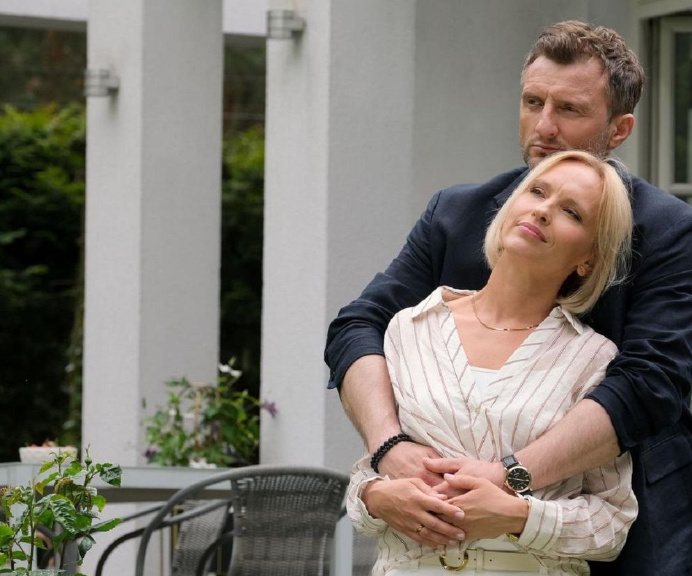 Dorota (Iwona Rejzner) i Bartek (Arkadiusz Smoleński) z M jak miłość przytulają się czule, stojąc w ogrodzie przed domem. Dorota, w jasnej koszuli w paski, z przymkniętymi oczami, opiera głowę na ramieniu Bartka, który obejmuje ją od tyłu. Ta scena odzwierciedla ich radość z rozpoczęcia procesu adopcyjnego, o czym przeczytasz więcej na Super Seriale.