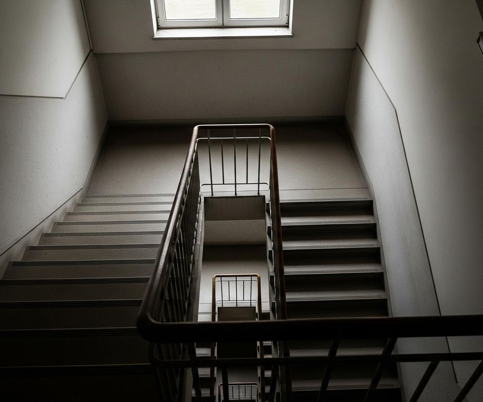 W kadrze znajduje się klatka schodowa widziana z góry, z perspektywy skierowanej w dół. Po obu stronach widoczne są biegi schodów z ciemnymi stopniami i jasnymi podstopnicami, rozdzielone balustradą z metalowymi pionowymi prętami i drewnianą poręczą w kolorze brązowym. Na szczycie klatki schodowej, pośrodku kadru, umieszczone jest okno z białymi ramami, przez które wpada naturalne światło, rozświetlając część sufitu i ścian. Dominują szare i ciemne odcienie, z wyraźnymi kontrastami światła i cienia, tworzącymi geometryczne kształty na ścianach.