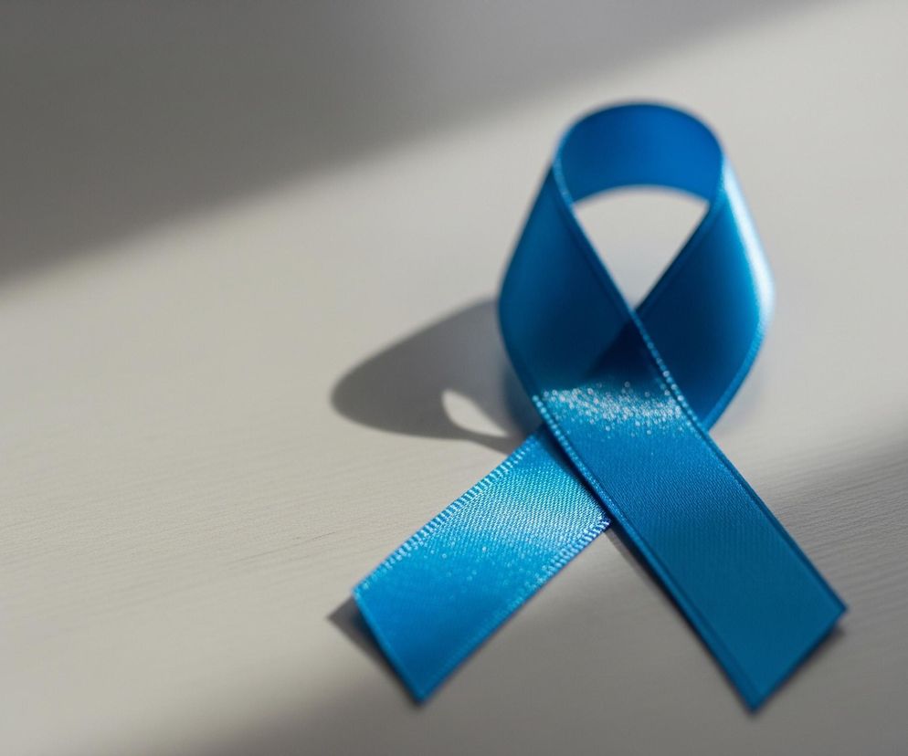 Niebieska wstążka satynowa uformowana w pętlę i skrzyżowanie, symbolizujące świadomość, leży na jasnej, płaskiej powierzchni. Po prawej stronie wstążki, na górnym jej zagięciu, widać jaśniejszy odcień koloru niebieskiego i błysk światła. Cień wstążki jest wyraźnie widoczny na powierzchni, zwłaszcza za górną pętlą i pod skrzyżowanymi końcami. Tło stanowi szara powierzchnia, częściowo oświetlona, z widocznym, ukośnym pasmem światła przechodzącym przez lewą i środkową część kadru.