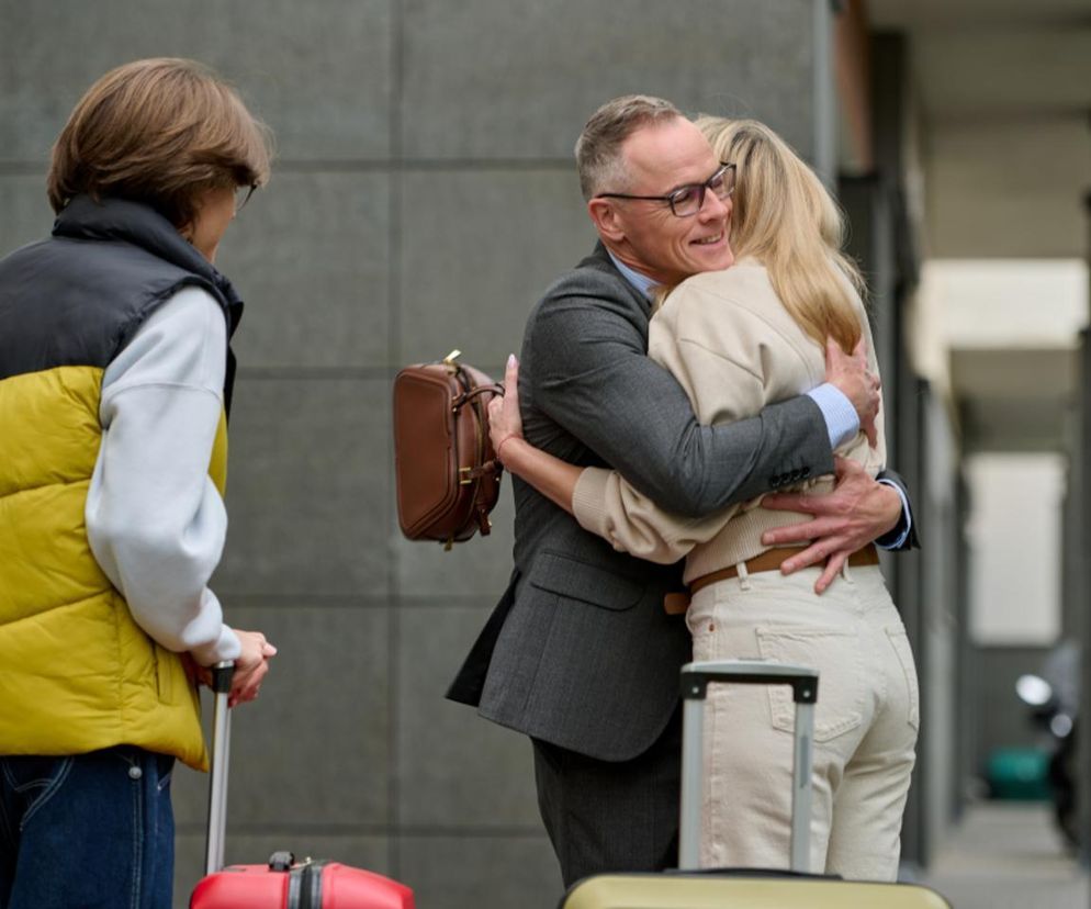 Damian Cieślik z serialu Na Wspólnej serdecznie obejmujący Monikę, oboje wyrażają radość z powrotu do domu po trudnych wydarzeniach. Obok stoi Kajtek z bagażami, co symbolizuje ich ponowne zjednoczenie. Cała rodzina jest szczęśliwa, wracając do normalności. Więcej na ten temat znajdziesz na Super Seriale.