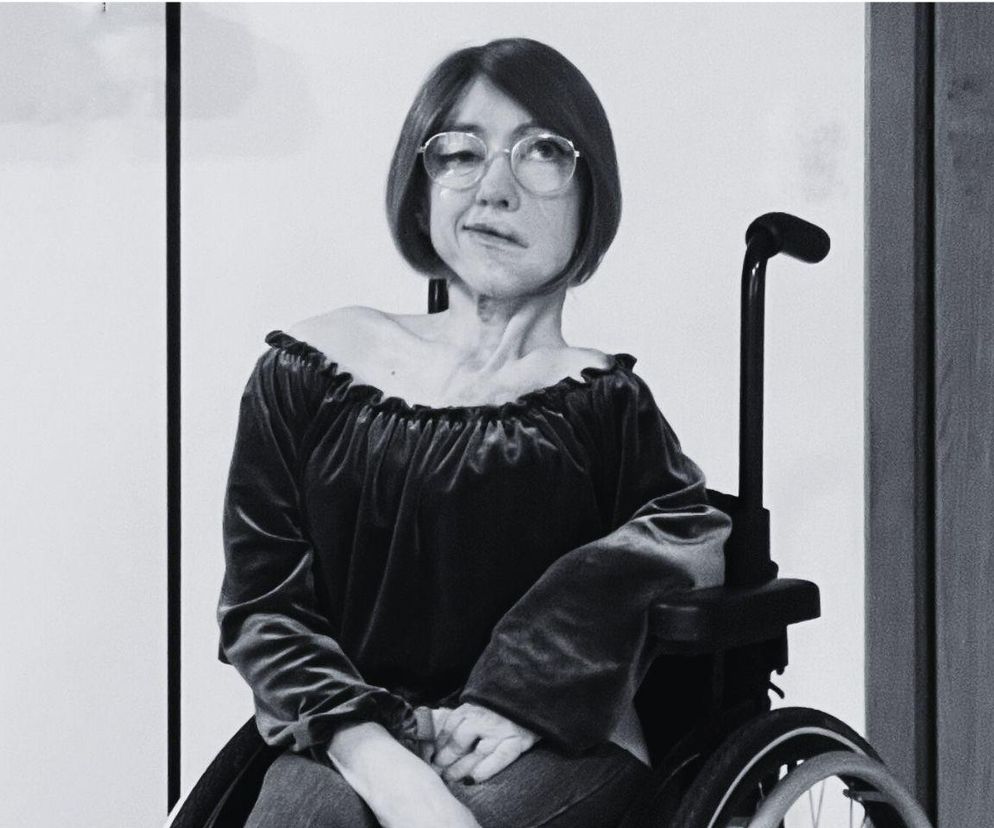 Katarzyna Wolińska, influencerka i autorka profilu „Pudel i ja”, siedzi na wózku inwalidzkim, ubrana w sukienkę z hiszpańskim dekoltem. Czarno-białe zdjęcie podkreśla powagę tematu, jakim jest jej walka z rzadką chorobą i rakiem tarczycy, o czym przeczytasz więcej na Poradnik Zdrowie.