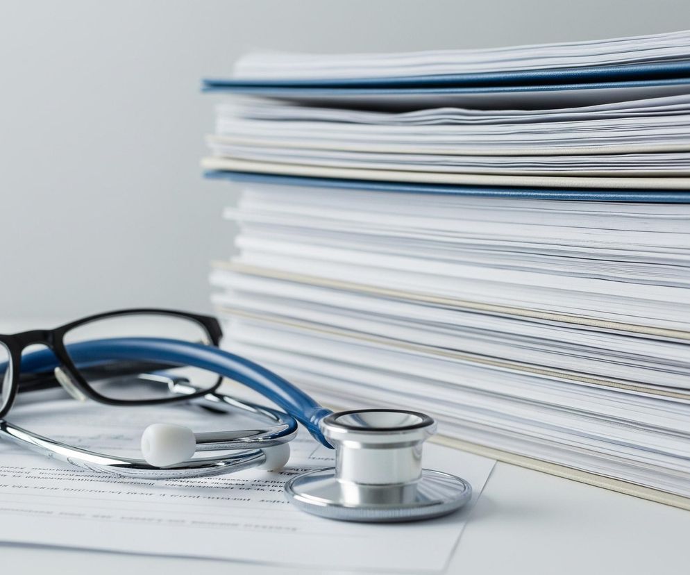 Na białym stole leżą okulary w czarnej oprawce oraz niebieski stetoskop z białymi oliwkami i srebrną głowicą. Przedmiotami tymi leżą na kartkach papieru z niewyraźnym tekstem. W tle, po prawej stronie, wznosi się wysoki stos dokumentów i teczek, przeplatanych niebieskimi i jasnobrązowymi okładkami, na tle jednolitej, jasnoszarej ściany. Główny plan jest ostry, podczas gdy tło jest lekko rozmyte.