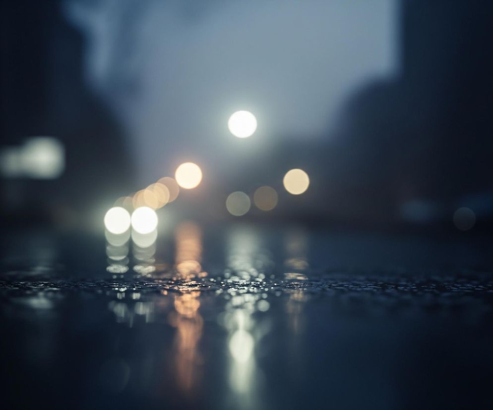 Droga pokryta jest drobnymi kroplami wody, które odbijają rozmyte światła w tle. W oddali widoczne są okrągłe, jasne plamy światła, głównie białe i żółtawe, ułożone w rząd sugerujący ulicę. Ciemne, rozmazane kształty po bokach i w górnej części obrazu wskazują na obiekty miejskie lub roślinność. Ogólny nastrój jest wieczorowy lub nocny, z dominującymi odcieniami niebieskiego, szarego i czarnego.