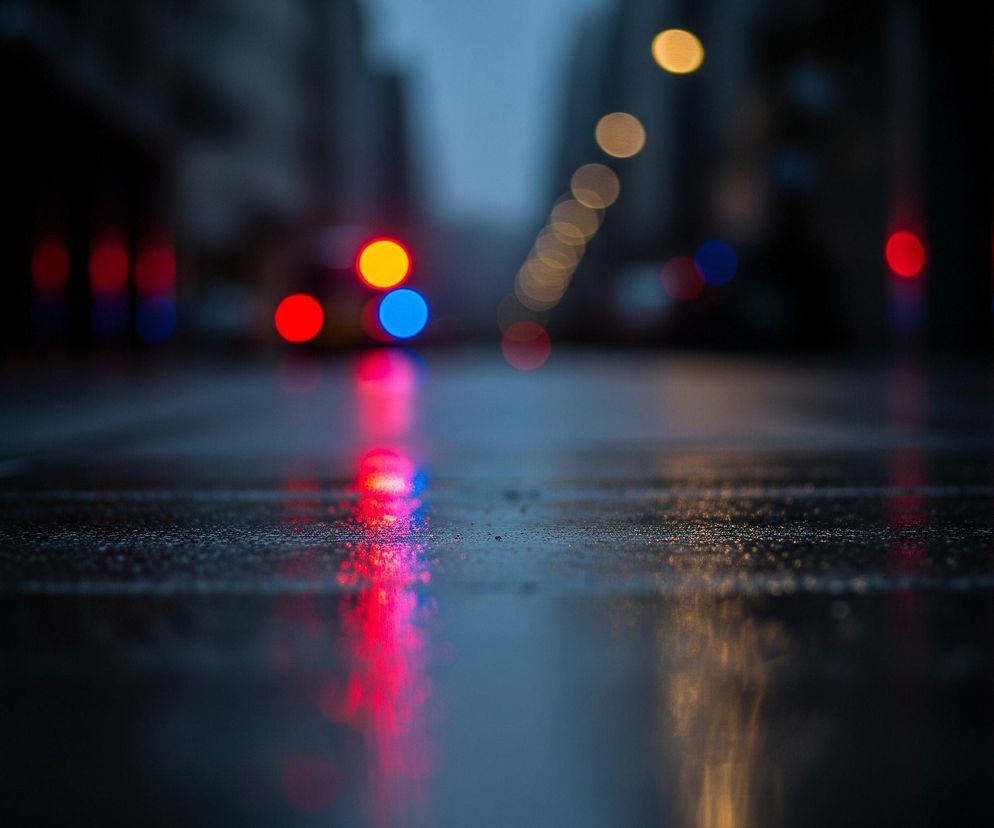 Mokra nawierzchnia, prawdopodobnie asfalt, pokryta jest drobnymi kroplami wody, odbijającymi jaskrawe, rozmyte światła. Główne, rozmazane plamy światła to czerwone, niebieskie i pomarańczowe okręgi, tworzące efekt bokeh. Światła te rozciągają się w dół, tworząc pionowe smugi odbite w wilgotnej powierzchni. W tle widoczne są niewyraźne zarysy budynków i ulicy, pogrążone w ciemności.
