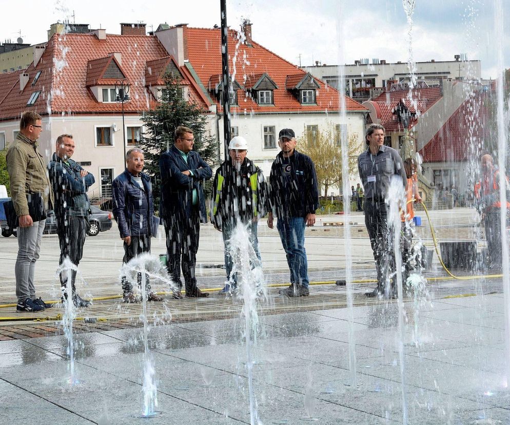 Grupa osób, w tym pracownicy i urzędnicy, testuje uruchomienie fontanny posadzkowej na Rynku w Myślenicach, gdzie woda tryska bezpośrednio z nawierzchni. Zdjęcie ilustruje wyzwania związane z nowoczesnymi rozwiązaniami w architekturze miejskiej, o których przeczytasz na Architektura Murator Plus.