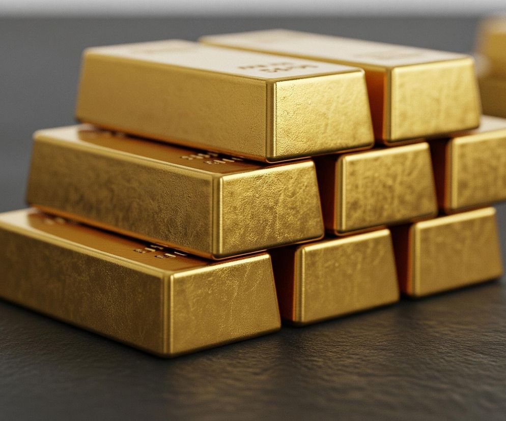 Na ciemnoszarym blacie leży stos sztabek złota. W pierwszym rzędzie widoczne są trzy sztabki, na nich leżą trzy kolejne, a na samej górze dwie. Sztabki mają matową, lekko chropowatą teksturę i metaliczny, złotawy połysk, z widocznymi refleksami światła. W tle, po prawej stronie, widać rozmyte, również złote obiekty, sugerujące obecność kolejnych sztabek.