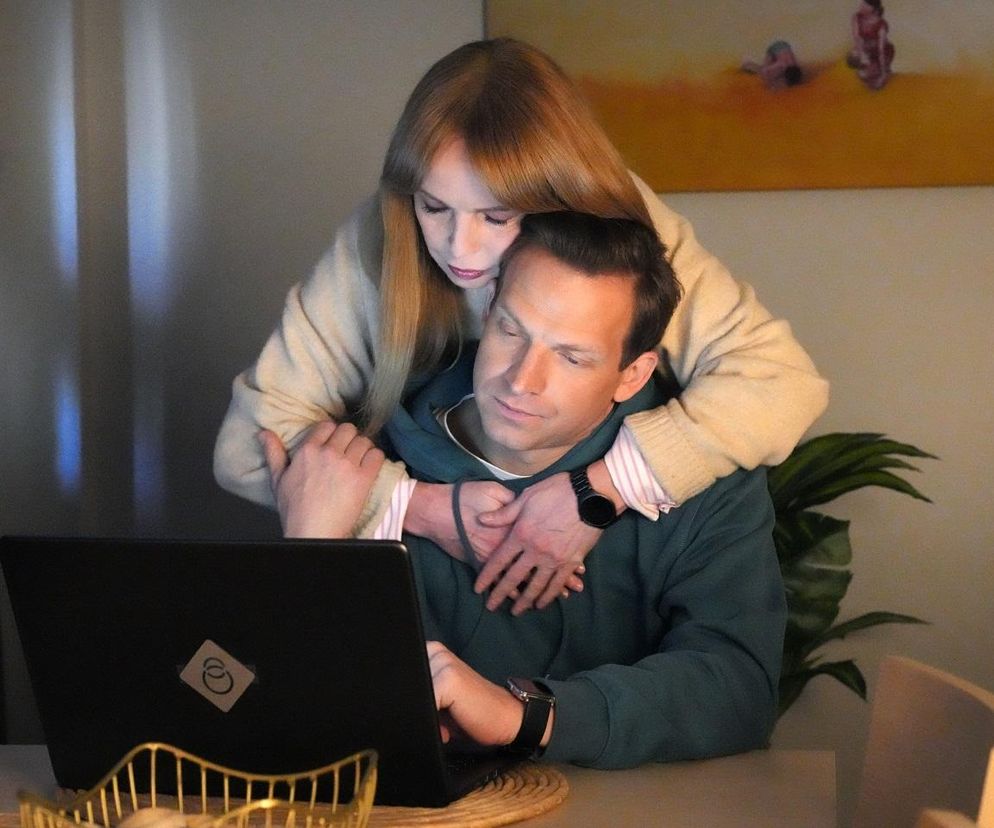 Łukasz Sadowski (Michał Rolnicki) w ciemnozielonej bluzie z kapturem pracuje na laptopie, a od tyłu obejmuje go Agnieszka (Katarzyna Tlałka) w beżowym swetrze, zaniepokojona. Ta scena to dramatyczny początek nowego życia Łukasza po zwolnieniu z Szoku, o czym przeczytasz więcej na Super Seriale.