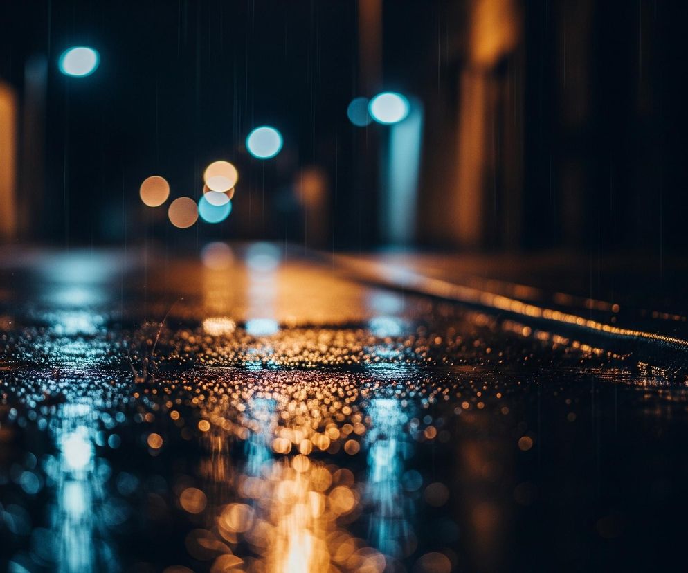 Wilgotna powierzchnia, prawdopodobnie asfalt ulicy, wypełnia dolną część kadru, pokryta drobnymi kroplami wody odbijającymi światła. Rozmyte punkty świetlne o barwach pomarańczowych i niebiesko-zielonych rozświetlają głębię obrazu, sugerując nocne otoczenie miasta. Pionowe, cienkie linie widoczne w górnej części kadru świadczą o padającym deszczu.