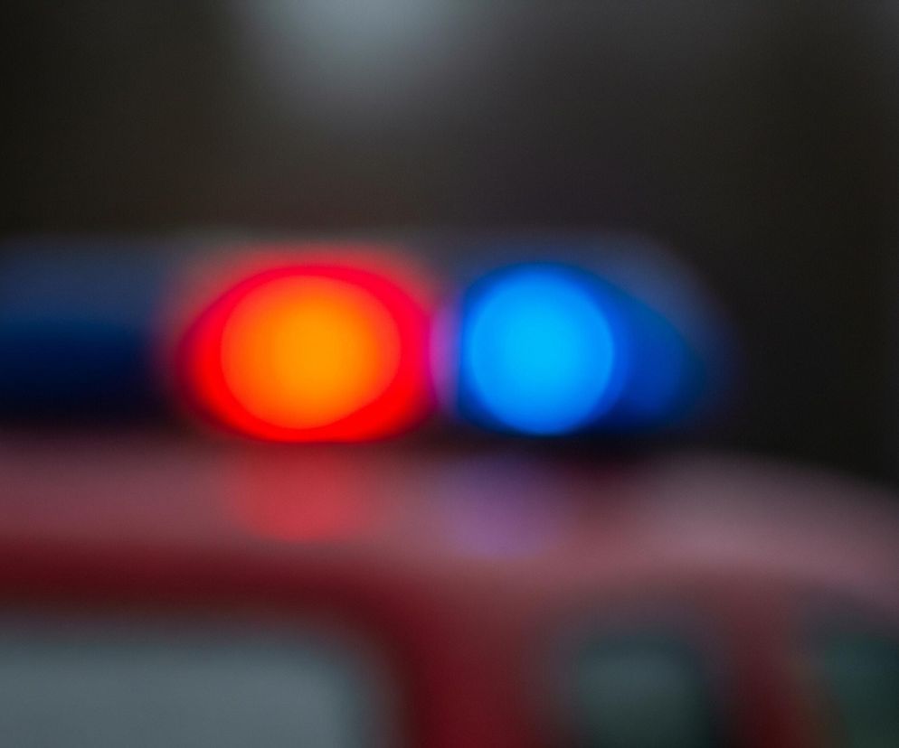 Na pierwszym planie, centralnie, widoczne są dwa rozmyte, intensywnie świecące okręgi: czerwony po lewej i niebieski po prawej, osadzone na ciemnym tle. Poniżej świateł znajduje się rozmyta czerwona powierzchnia, sugerująca karoserię pojazdu. Tło jest ciemne i zamazane, z niewielkimi jaśniejszymi smugami u góry obrazu.