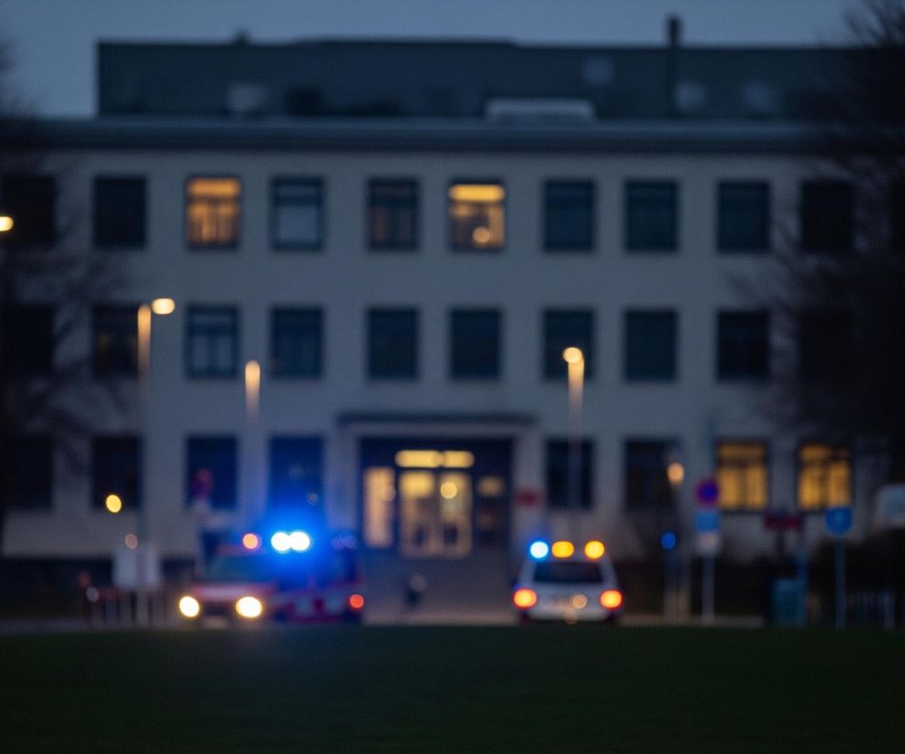 Centralnie widoczny jest rozmazany, trzypiętrowy budynek z licznymi oknami, niektóre z nich świecą żółtym światłem. Przed budynkiem, na placu, stoją dwa pojazdy z włączonymi światłami awaryjnymi: po lewej widać czerwono-biały samochód z niebieskimi światłami sygnalizacyjnymi na dachu, a po prawej biały samochód z żółtymi i niebieskimi światłami. Scena jest słabo oświetlona, panuje zmierzch lub noc, a na pierwszym planie znajduje się ciemnozielony trawnik. Wzdłuż placu rozmieszczone są latarnie uliczne emitujące ciepłe, żółte światło.