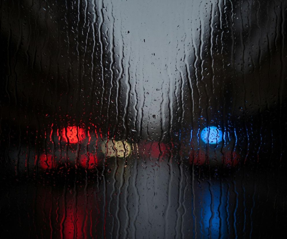 Szklana powierzchnia, prawdopodobnie szyba, pokryta jest licznymi kroplami deszczu i smugami wody, które spływają pionowo. W tle, za zamgloną i mokrą szybą, widać rozmyte, kolorowe światła. Dominują światła czerwone po lewej stronie i niebieskie po prawej, a także pojedyncze, żółte światło w centrum. Całe tło jest ciemne, prawie czarne, z wyjątkiem jasnych plam świateł, co tworzy efekt bokeh.