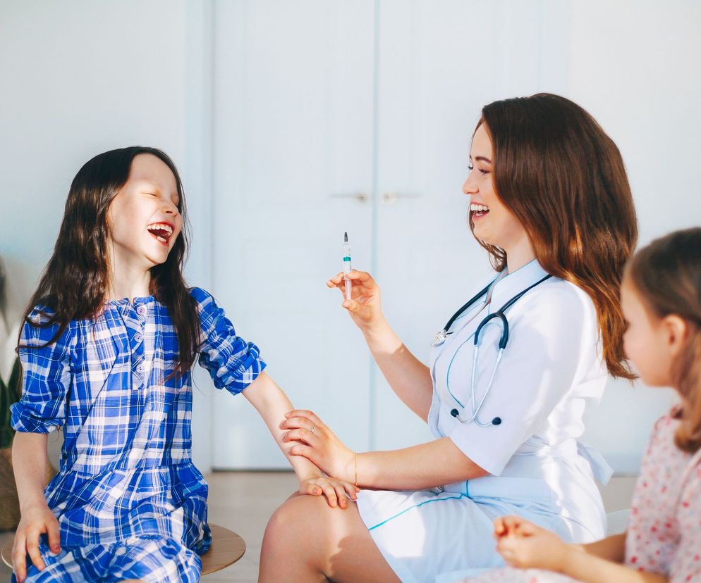 Uśmiechnięta pielęgniarka w białym kitlu i ze stetoskopem przygotowuje strzykawkę do szczepienia dziewczynki w kratkowanej sukience, która śmieje się głośno. Obok siedzi drugie dziecko. W tle jasne wnętrze z rośliną. To zdjęcie ilustruje bezpieczeństwo szczepień u dzieci, o czym przeczytasz więcej na Poradnik Zdrowie.