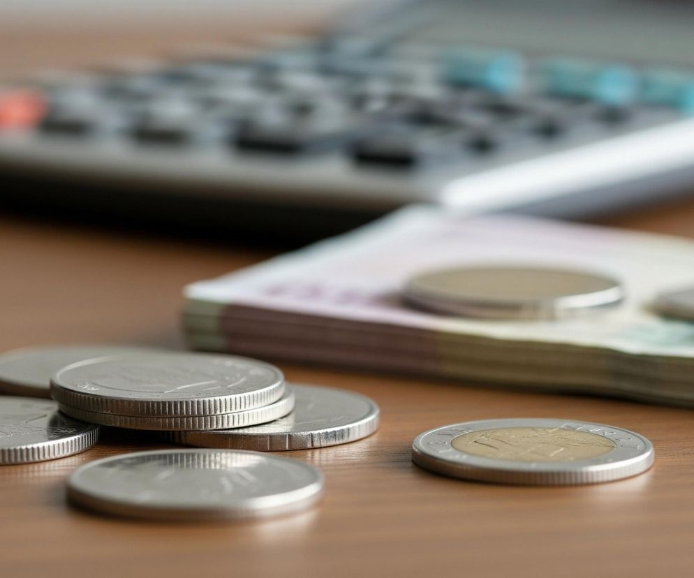 Monety i banknoty obok kalkulatora na drewnianym stole symbolizują rozliczenia podatkowe i finanse firmy, o których szerzej przeczytasz na Super Biznes. Zdjęcie ilustruje zmiany w kasach fiskalnych i sposoby unikania obowiązku ich posiadania.