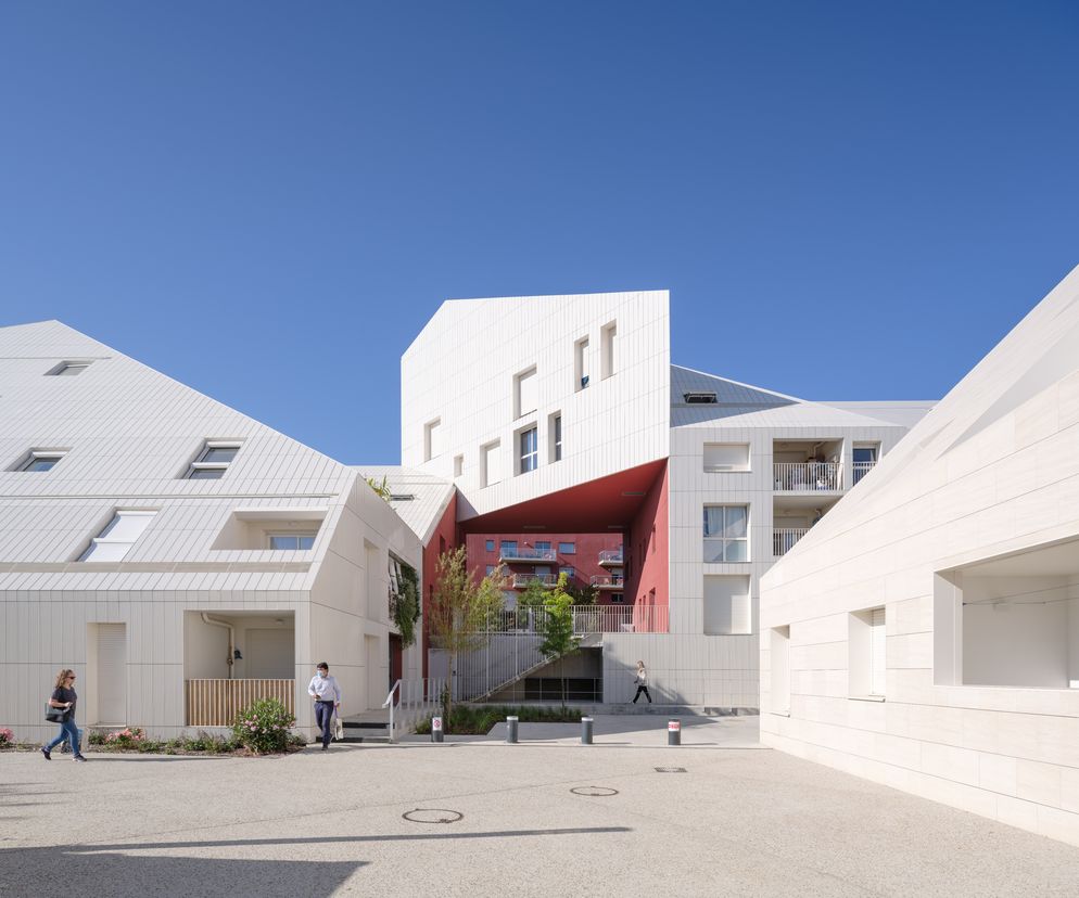 Nowoczesny kompleks apartamentowców Ilot Queyries w Bordeaux z białymi elewacjami i spadzistymi dachami, z widocznym czerwonym wnętrzem portalu. Na placu przemieszczają się ludzie, a o kompleksie i architekturze MVRDV przeczytasz na Architektura Murator Plus.