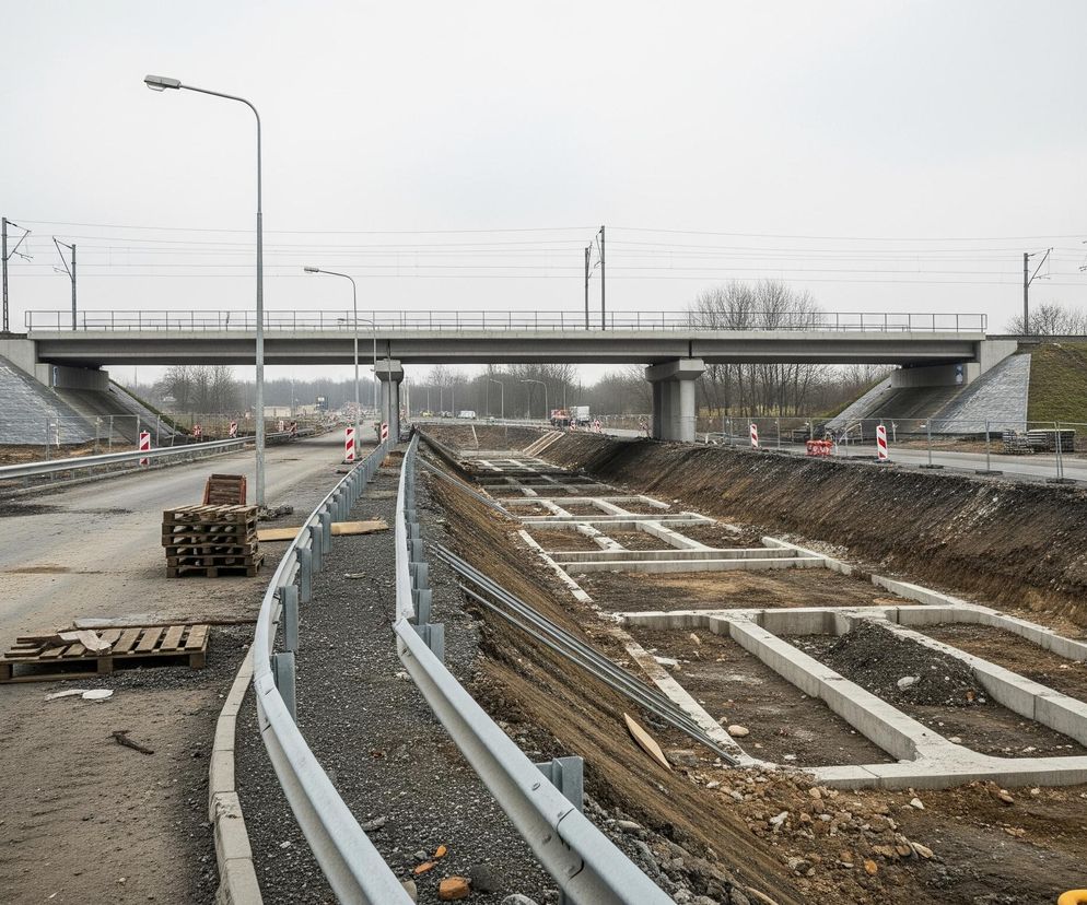 Na zdjęciu widoczna jest budowa drogi pod wiaduktem kolejowym w szary dzień. Na pierwszym planie, po lewej stronie, biegnie asfaltowa droga w trakcie budowy, z poboczem i leżącymi paletami, a po prawej stronie znajduje się głęboki wykop z betonowymi fundamentami. Wzdłuż drogi i wykopu ustawione są metalowe barierki ochronne oraz biało-czerwone słupki drogowe. W tle, nad drogą i wykopem, rozciąga się wiadukt kolejowy z dwoma torami i trakcją elektryczną, wsparty na betonowych filarach i otoczony zielonymi nasypami.