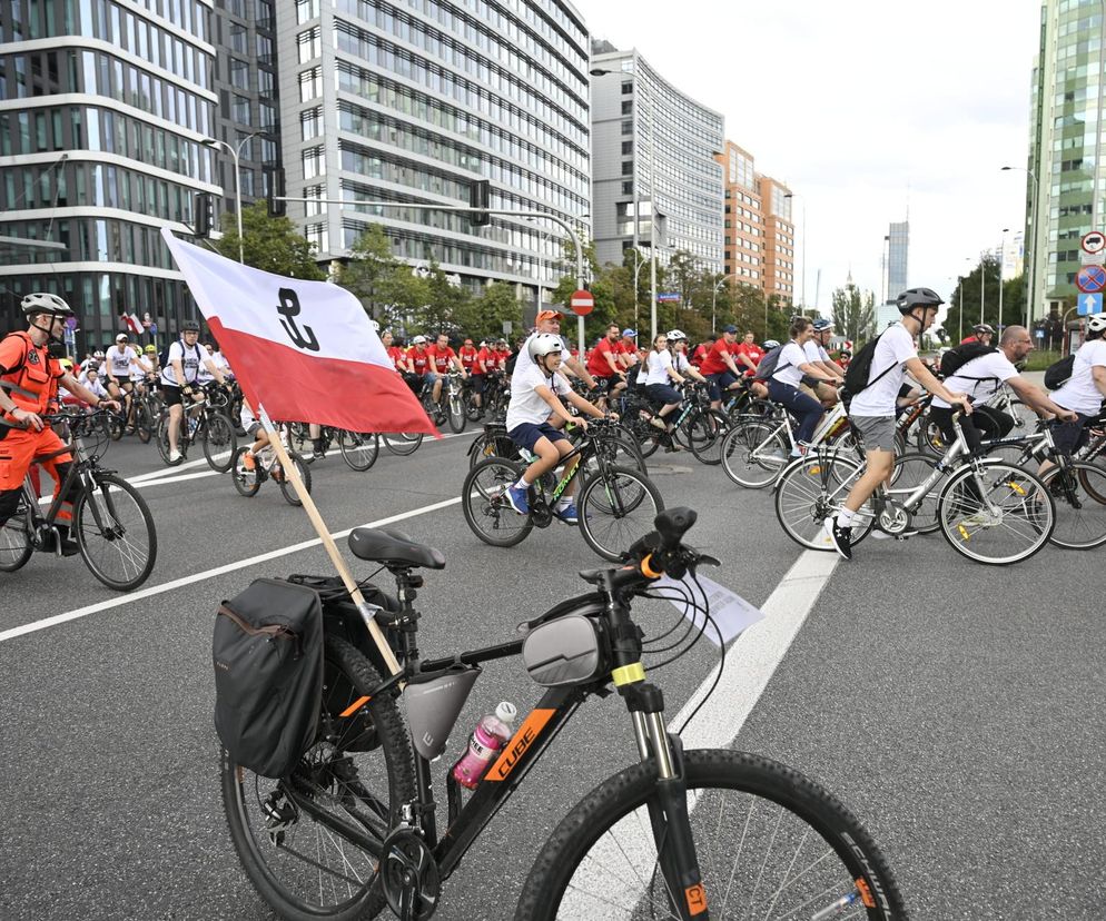 Peleton rowerzystów w białych i czerwonych koszulkach z biało-czerwoną flagą na pierwszym planie, podczas przejazdu upamiętniającego Powstanie Warszawskie. Grupa rowerzystów na ulicach miasta symbolizuje pamięć historyczną, o czym przeczytasz na Super Biznes.